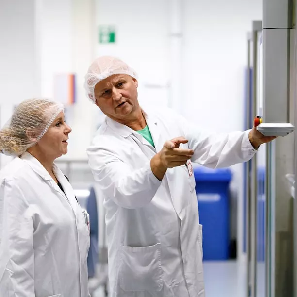 Two healthcare professionals in lab coats and hairnets engage in conversation. The man, gesturing towards a device, appears to be explaining something, while the woman listens attentively. The background features medical equipment and a waste bin.