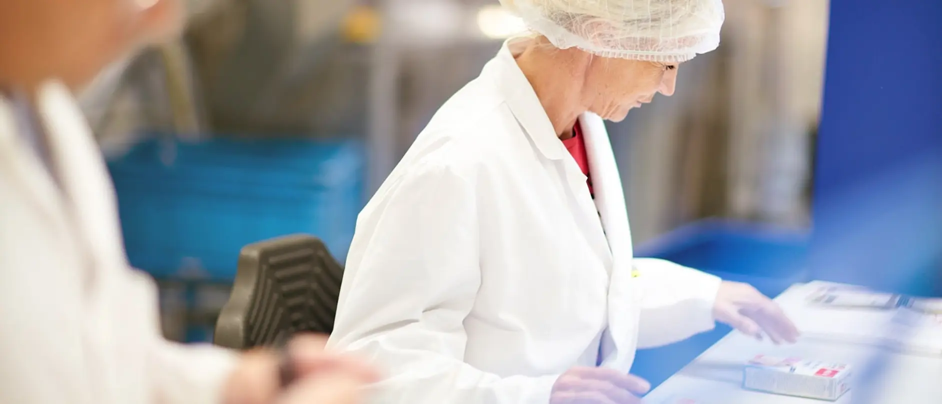 A woman in a lab coat and hairnet examines products on a table in a clean room environment. She appears focused on her work, ensuring accuracy in a professional setting, possibly related to pharmaceuticals or quality control.