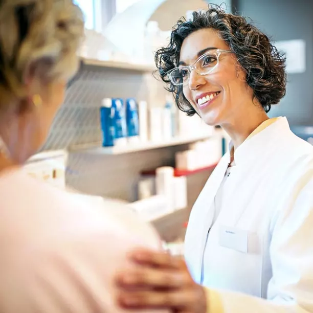 A smiling healthcare professional in a white coat is engaging warmly with a patient in a pharmacy. Shelves in the background display various health and beauty products, creating a welcoming atmosphere for consultation and care.