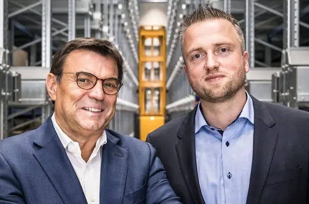 Two men are posing for a photo in a warehouse setting, standing in front of shelving units. One man is wearing glasses and a suit, smiling confidently, while the other is in a dress shirt and jacket, looking serious. The background features organized storage shelves.