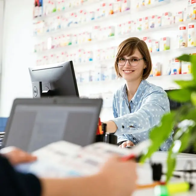 A smiling customer service representative in a blue polka-dot shirt sits at a desk, assisting a customer. In the background, a wall displays various products, and houseplants add a touch of greenery to the scene.