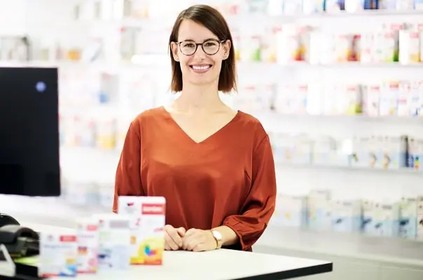 A smiling woman with short hair and glasses stands behind a counter in a pharmacy. She wears an orange blouse and is surrounded by shelves stocked with various medication boxes. In front of her, a few boxes of medicines are placed on the counter.