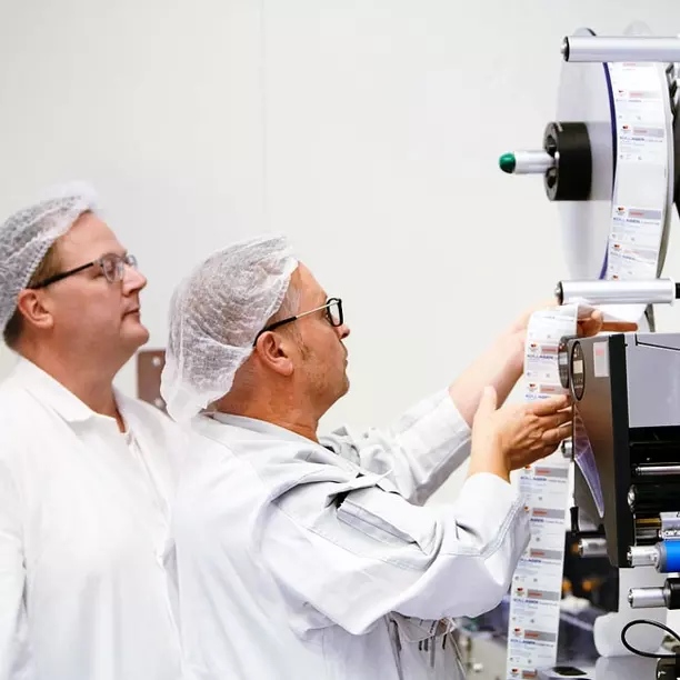 Two men in white lab coats and hairnets are working with a label printing machine. One man is carefully guiding a roll of printed labels, while the other observes, focused on the process. The setting appears to be a clean, industrial environment.