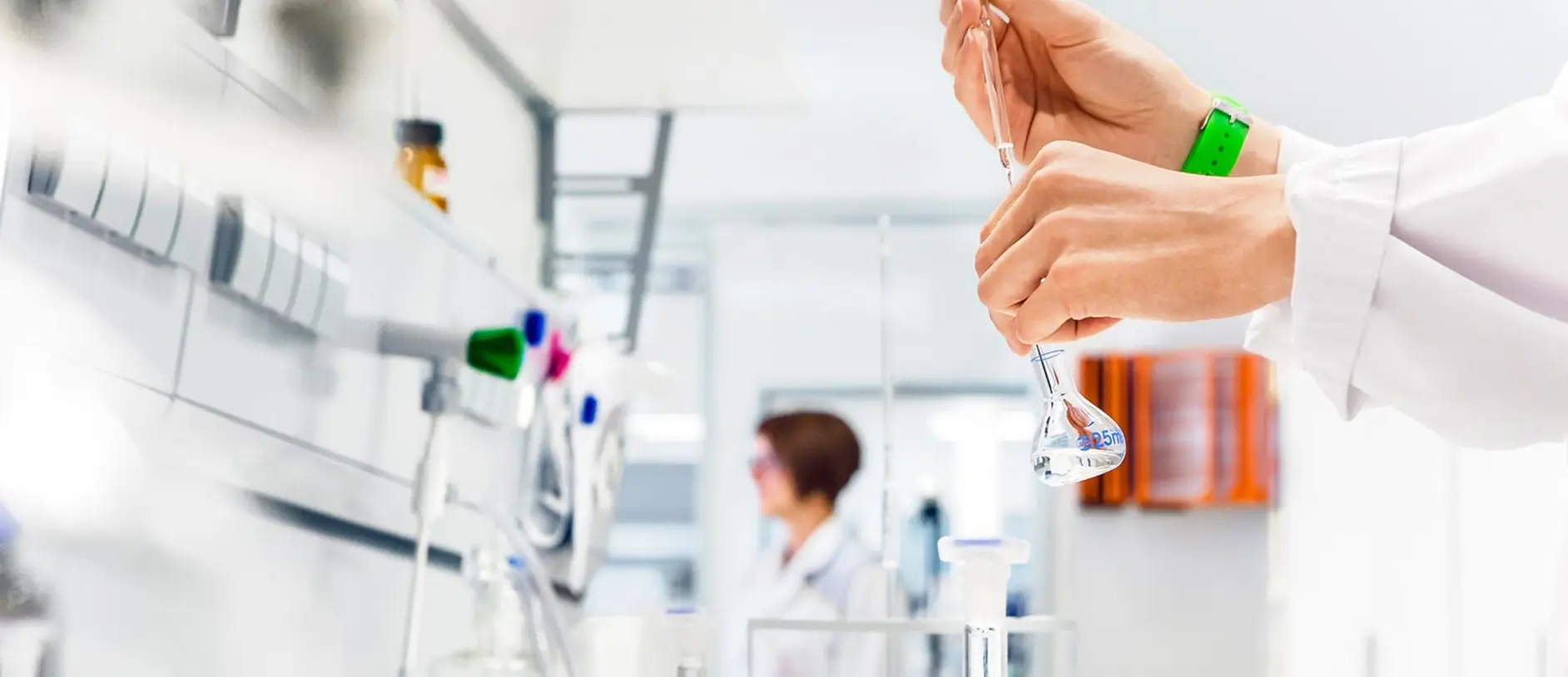 A laboratory scene with a person in a lab coat carefully holding a glass flask while preparing a solution. Another individual is visible in the background, indicating a collaborative workspace focused on scientific research. The setting is bright and organized, typical of a modern lab.