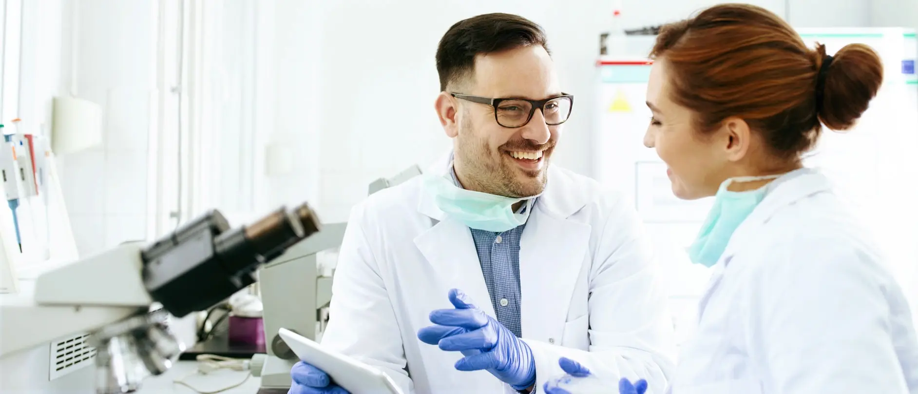 Two smiling laboratory professionals, a man and a woman, engage in a friendly conversation while wearing lab coats and face masks. The man holds a tablet, and they are surrounded by lab equipment and a bright, clean workspace, conveying a sense of collaboration and enthusiasm in their work.